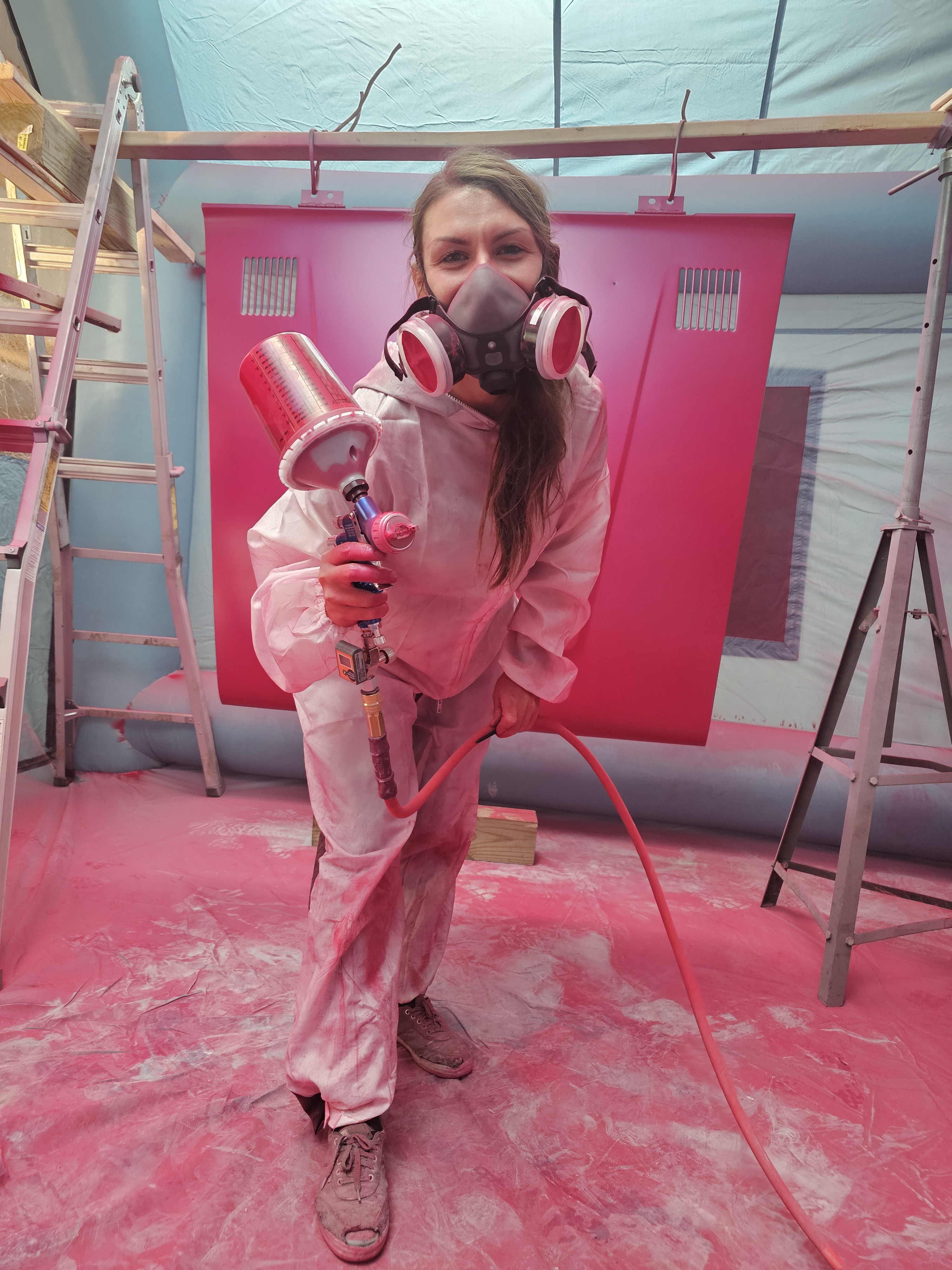 A woman wearing a protective suit holds a paint sprayer and is covered in hot pink paint.